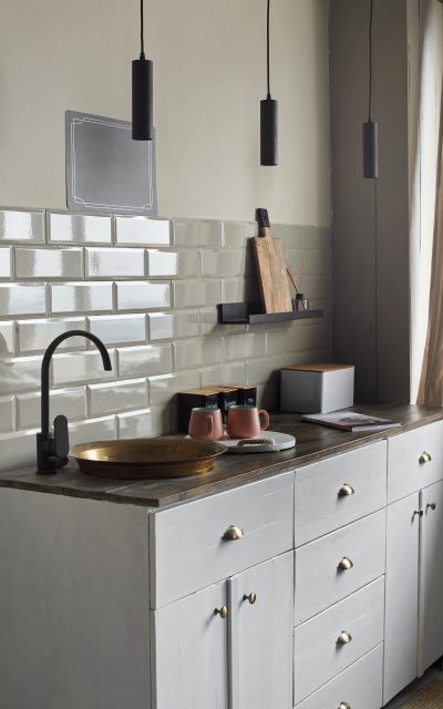 Modern creative idea of small kitchen with gray tiles wall with old fashion copper sink and modern faucet, tea and coffee containers, pink cups, and magazine on wooden counter.